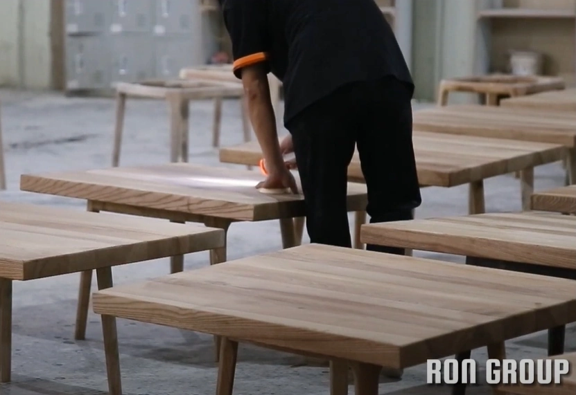 A quality assurance technician conducting a detailed surface inspection of a solid wood table on the Ron Group factory floor, ensuring product consistency.