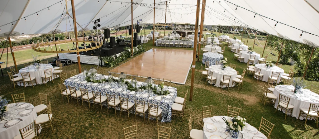 Large outdoor tent wedding reception featuring gold Chiavari chairs, long banquet tables, and a wooden dance floor.
