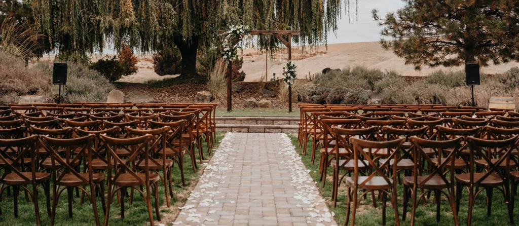 Rustic outdoor wedding ceremony setup featuring wooden cross-back chairs and a wooden arch under a large willow tree.