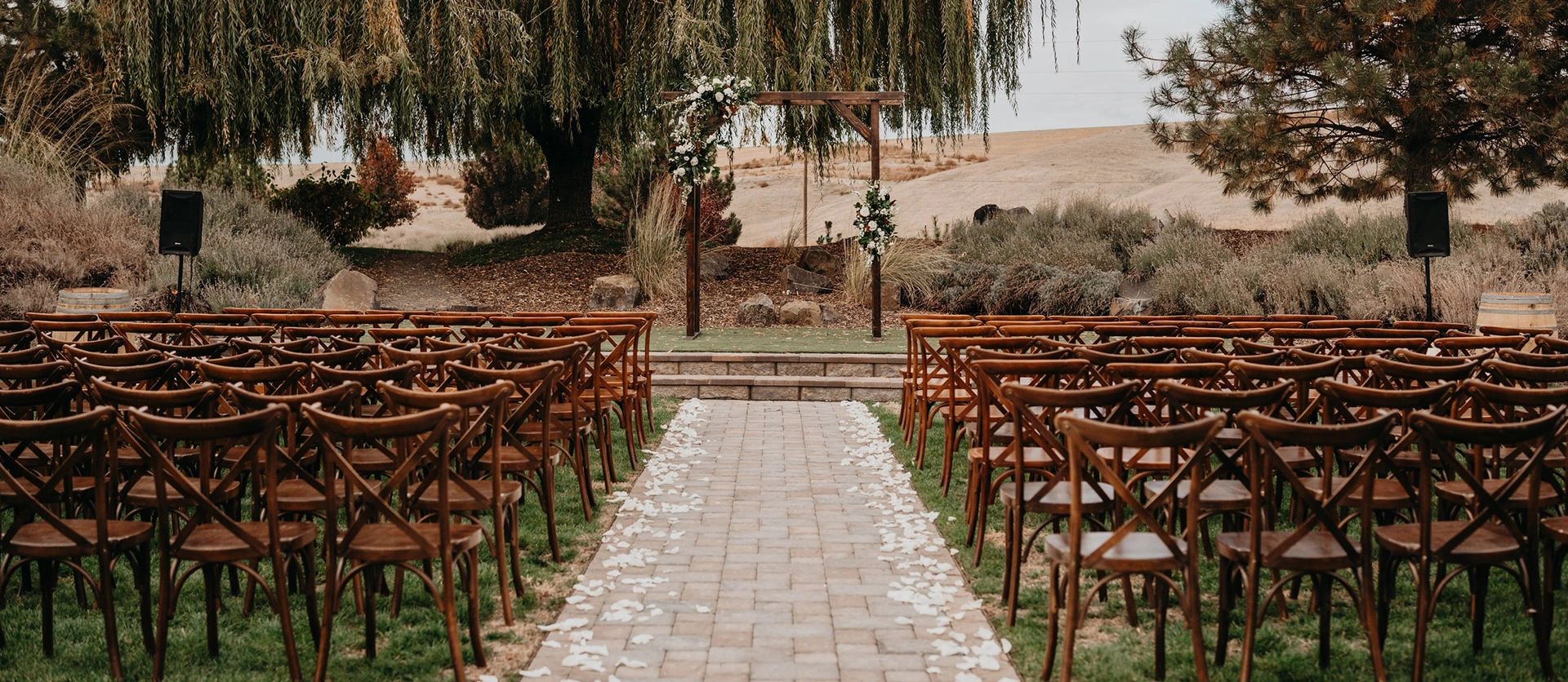 Rustic outdoor wedding ceremony setup featuring wooden cross-back chairs and a wooden arch under a large willow tree.